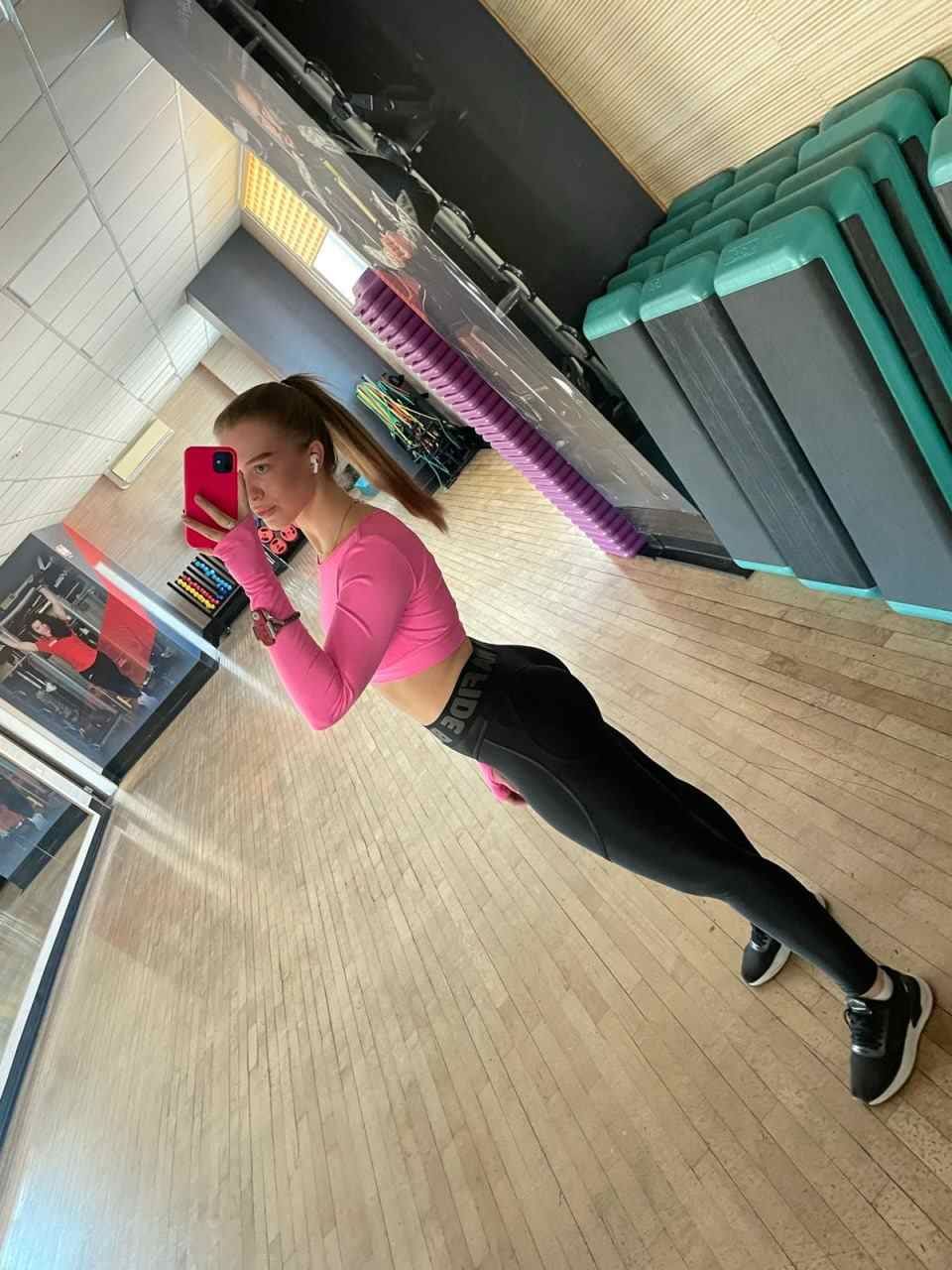 A woman with a ponytail stands in a gym holding a red phone, wearing a pink crop top, black leggings, and sneakers, with exercise mats and a mirror in the background.