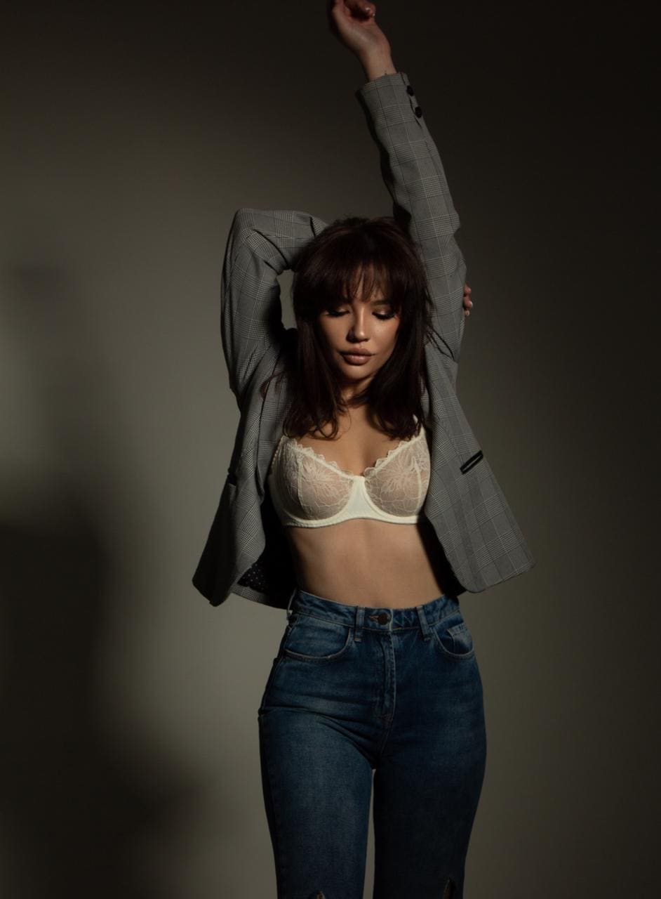 Woman with arms raised wearing an open grey plaid blazer over a white lace bra and blue jeans against a dark studio background.