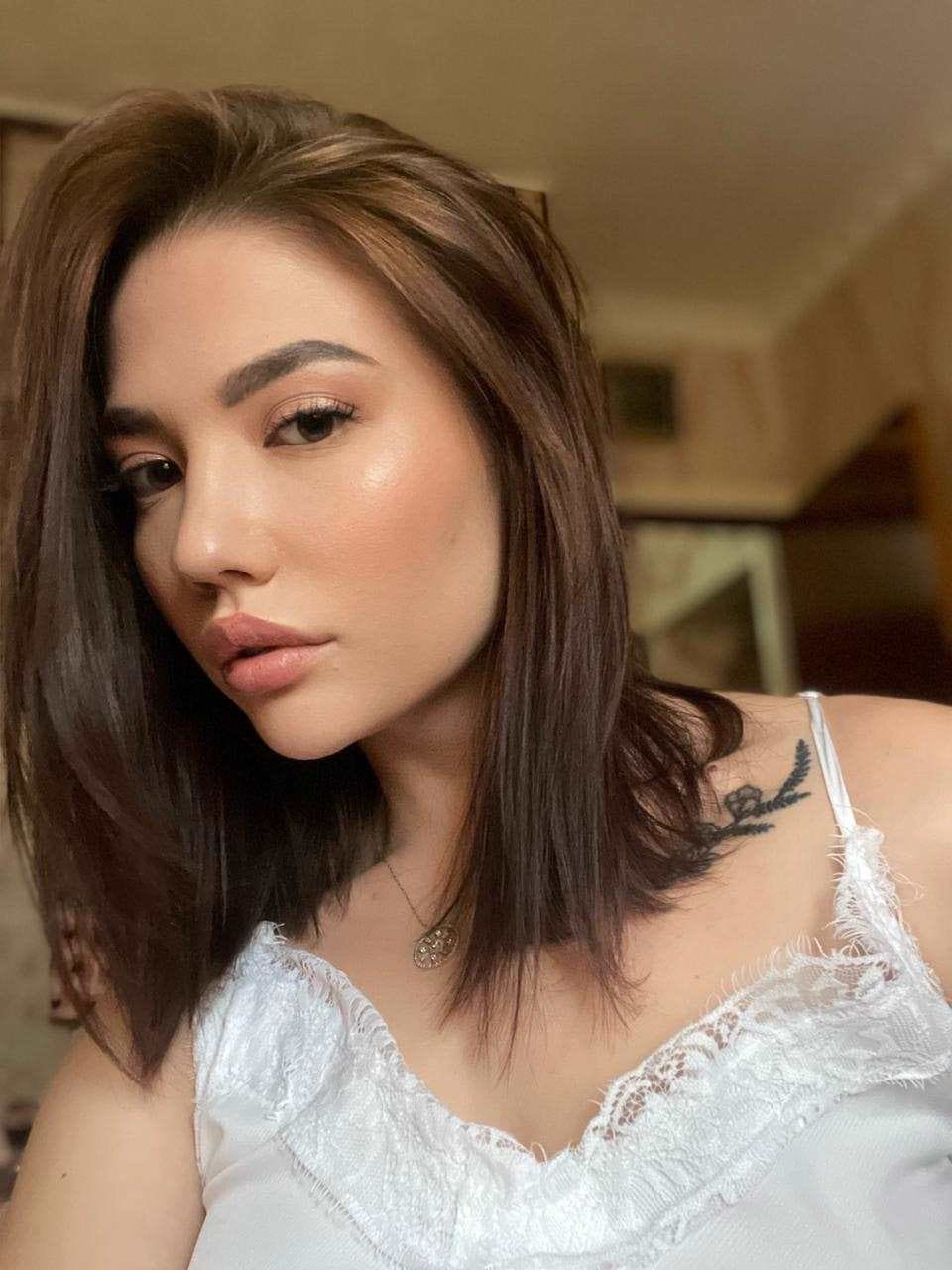 Alya in a close-up wearing a white lace-trimmed camisole and pendant necklace, with a floral tattoo on her left shoulder.