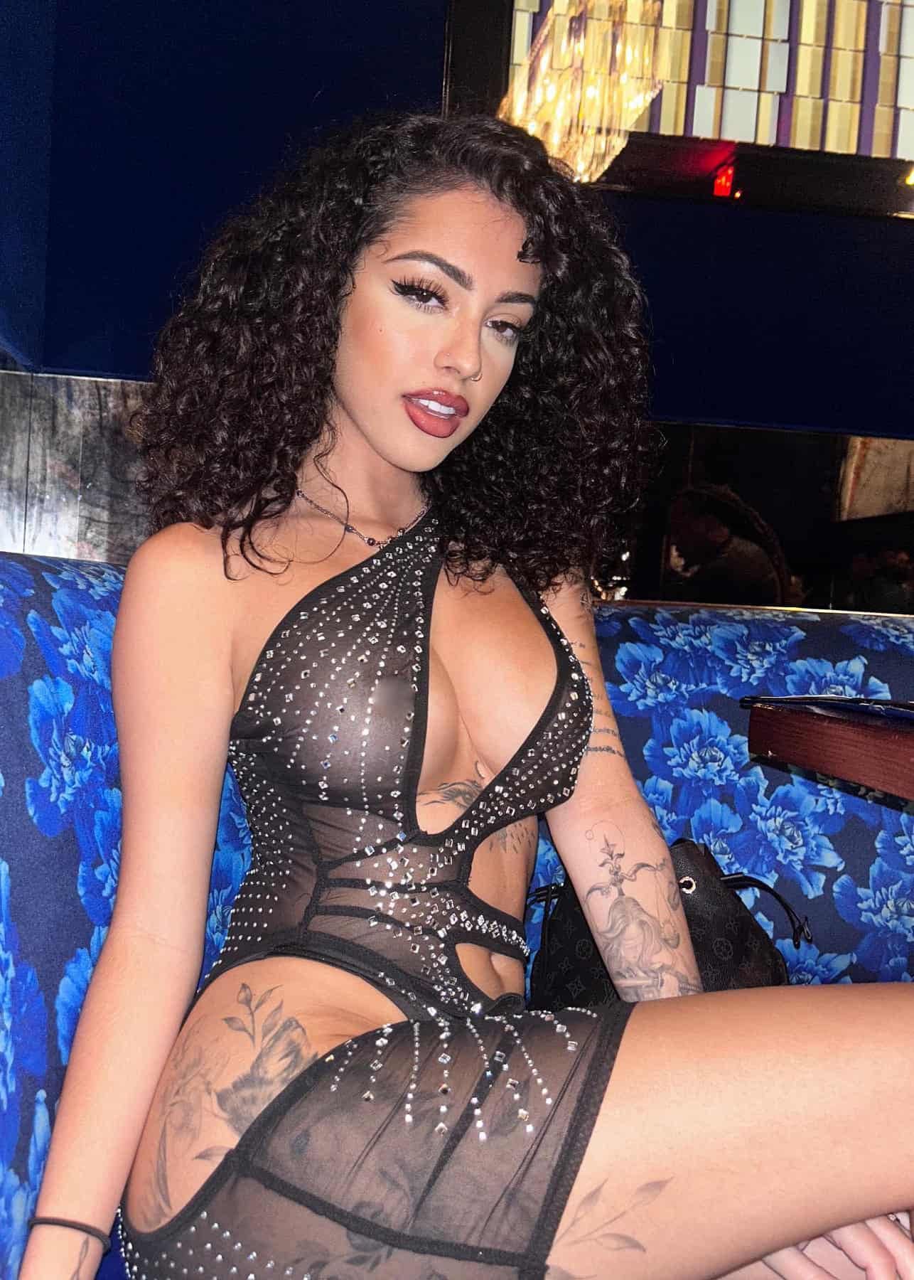 A woman with dark curly hair sits on a blue floral couch in a sheer black studded bodysuit, showing arm and hip tattoos.