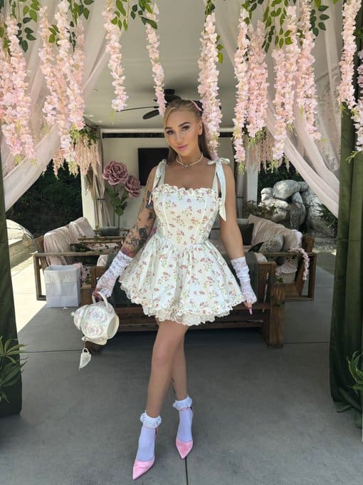 Eliana stands on a patio in a floral mini dress, white gloves, and pink heels, holding a handbag near hanging flowers.