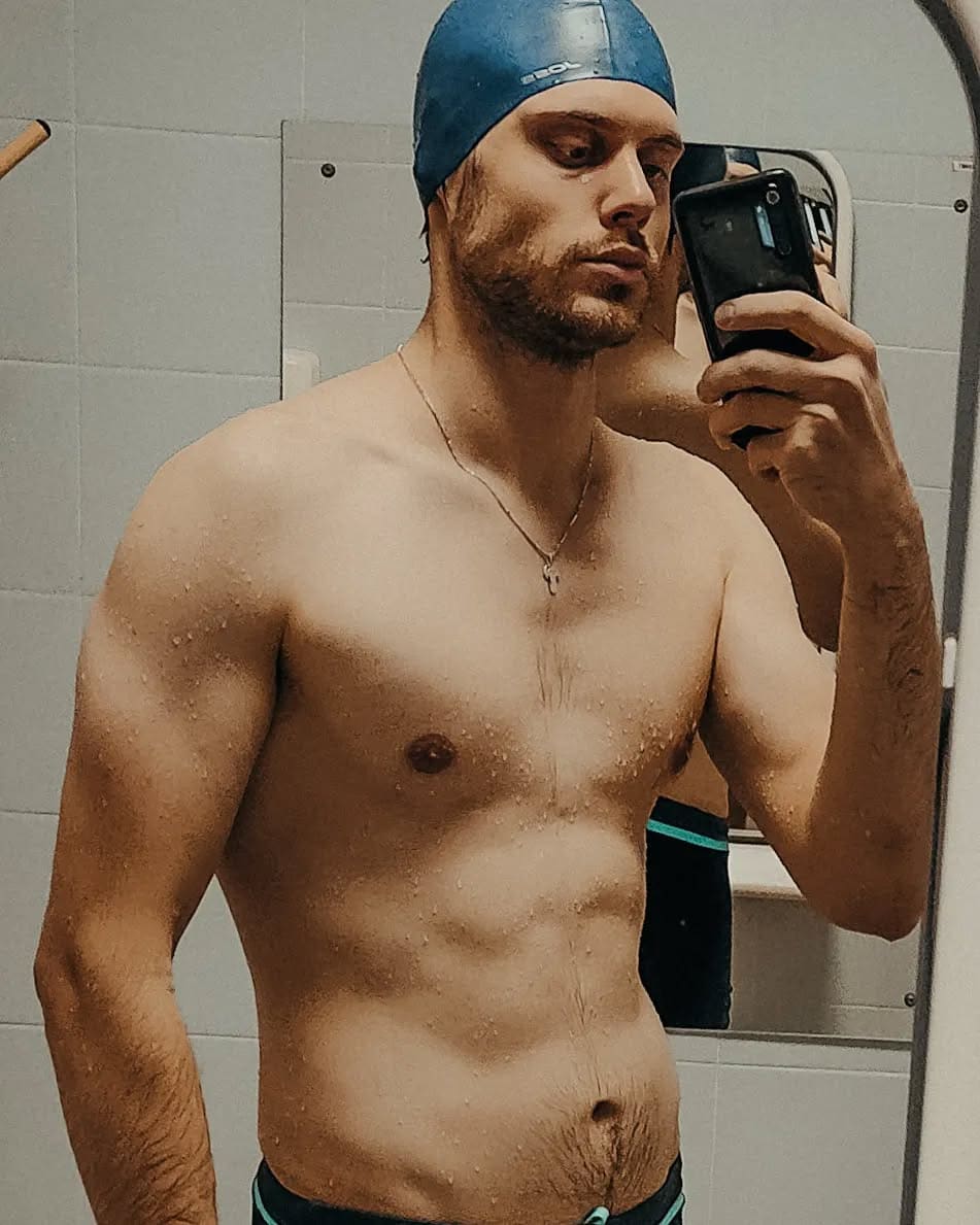A bearded man in a blue swim cap and dark trunks takes a mirror selfie with a black smartphone in a tiled room, skin appearing wet.