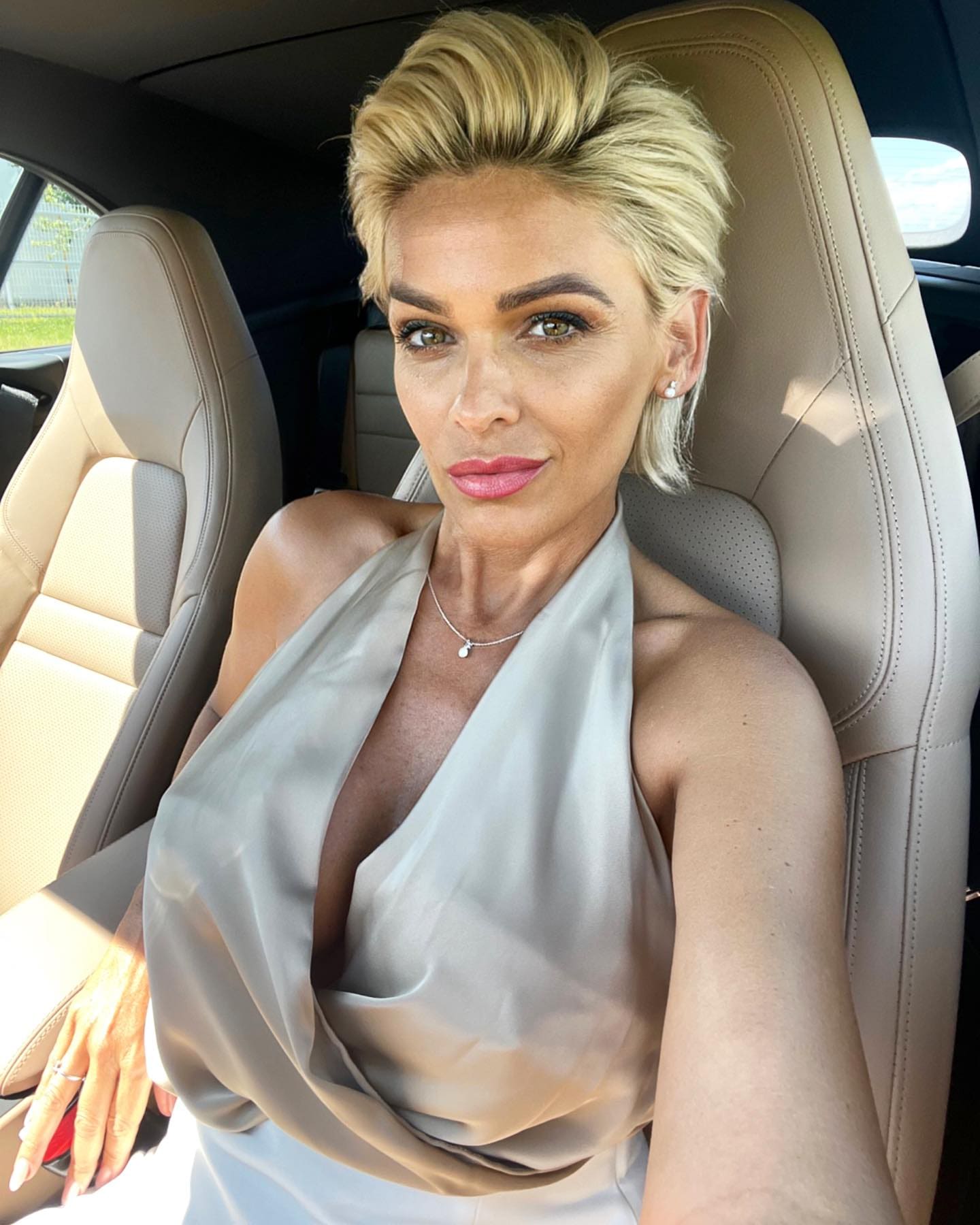 Adiza sits in a car driver's seat wearing a beige satin halter top, necklace, and pearls, with her hand on the steering wheel.