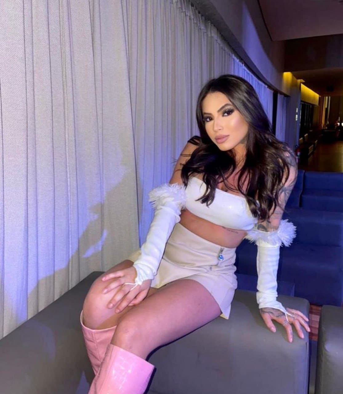 A woman sits on a gray bench wearing a white off-shoulder crop top with feathered sleeves, mini skirt, and pink boots.