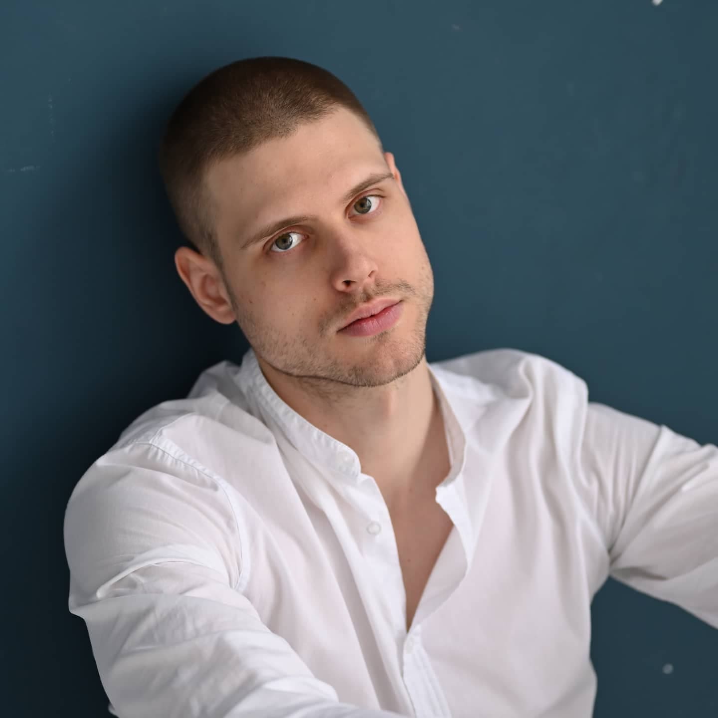 Miron leans against a dark teal wall, wearing an unbuttoned white shirt, looking toward the camera with a buzz cut and stubble.