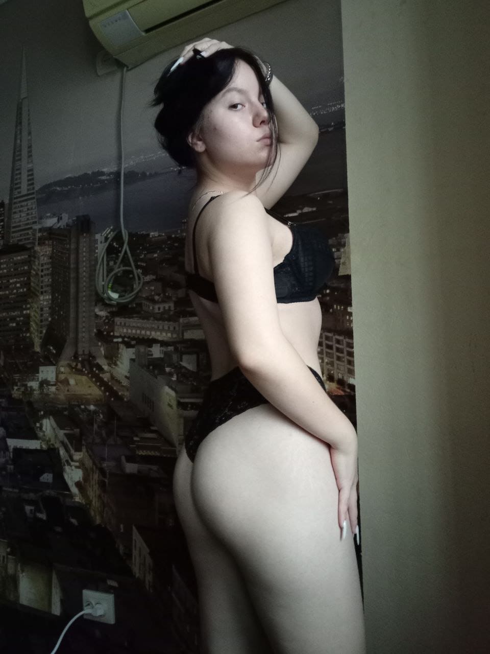 Woman with hair up in profile wearing black lace lingerie, hand raised to head, against a cityscape mural.