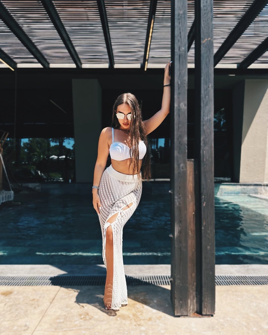 A woman in a white bikini top and long crochet skirt leans against a wooden post by a pool, wearing sunglasses.