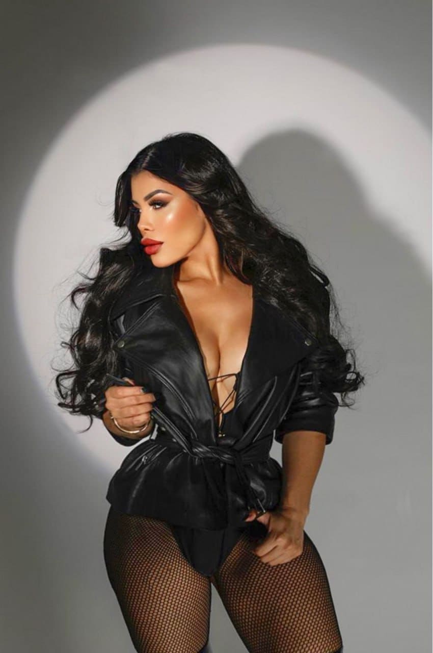 Woman standing in studio spotlight, wearing black leather jacket tied at waist over strappy top and fishnet tights, holding jacket open with one hand.