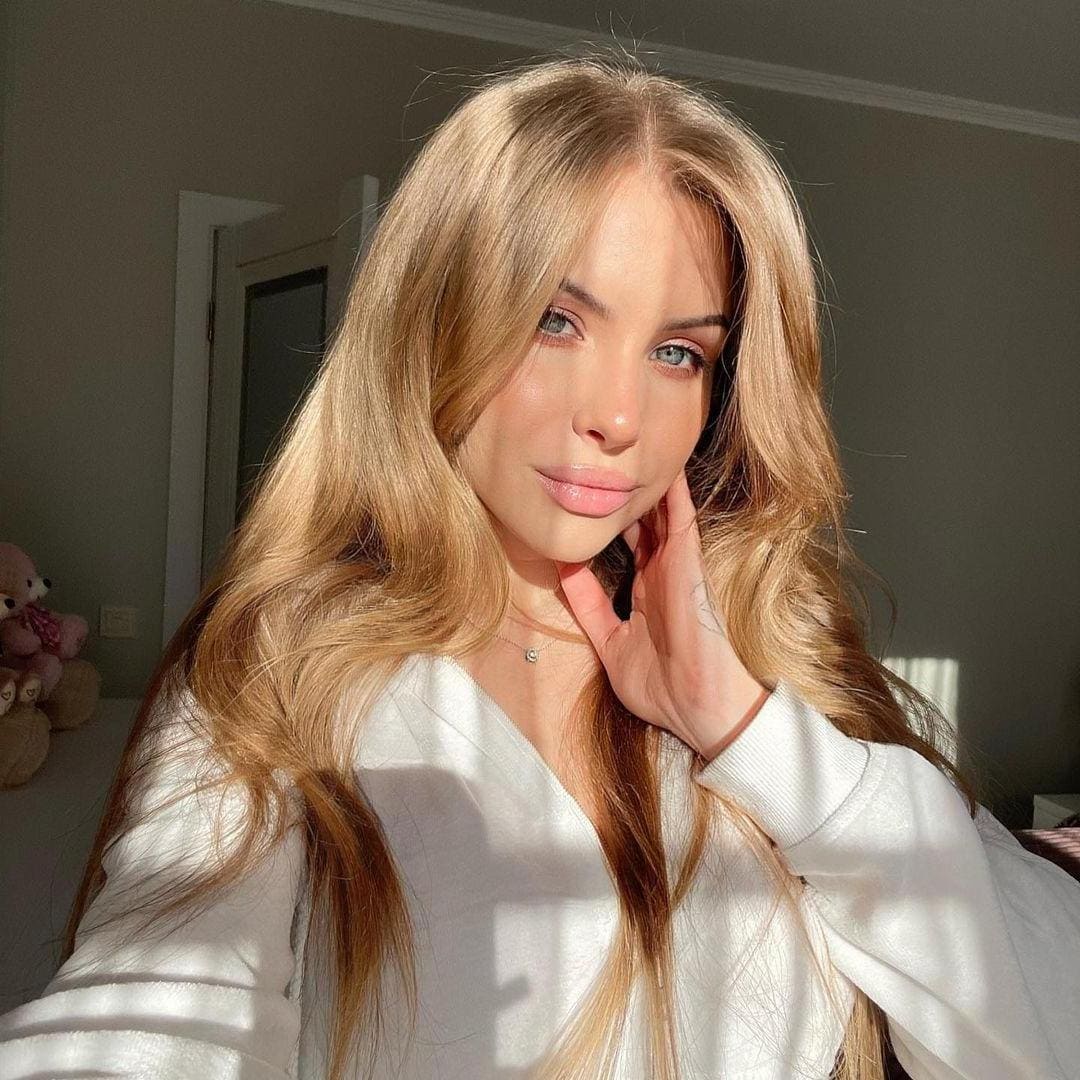 Hailey in a white long-sleeved top rests her hand on her cheek indoors, with sunlight shadows and stuffed toys in the background.