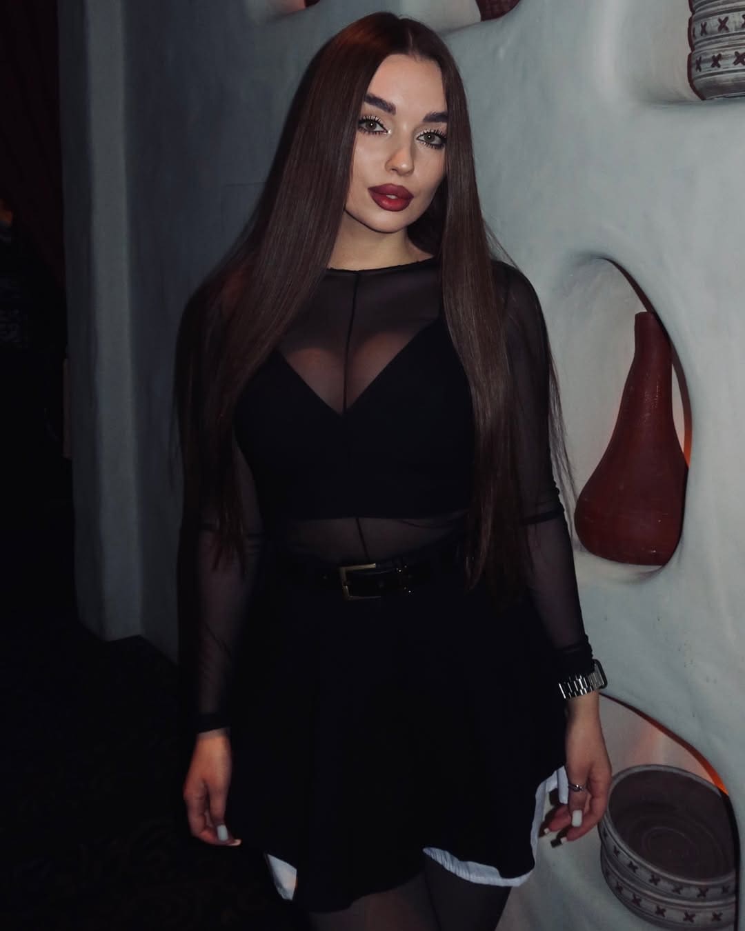A woman in a black dress with sheer sleeves and a belted waist stands facing the camera against a textured white wall.