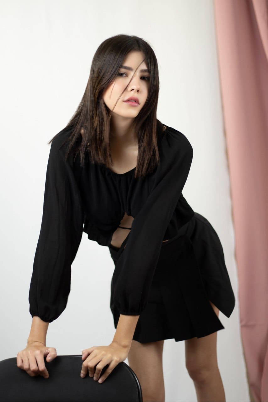 A woman leans on a black chair wearing a black cutout top and pleated mini skirt, against a white wall with a pink curtain.