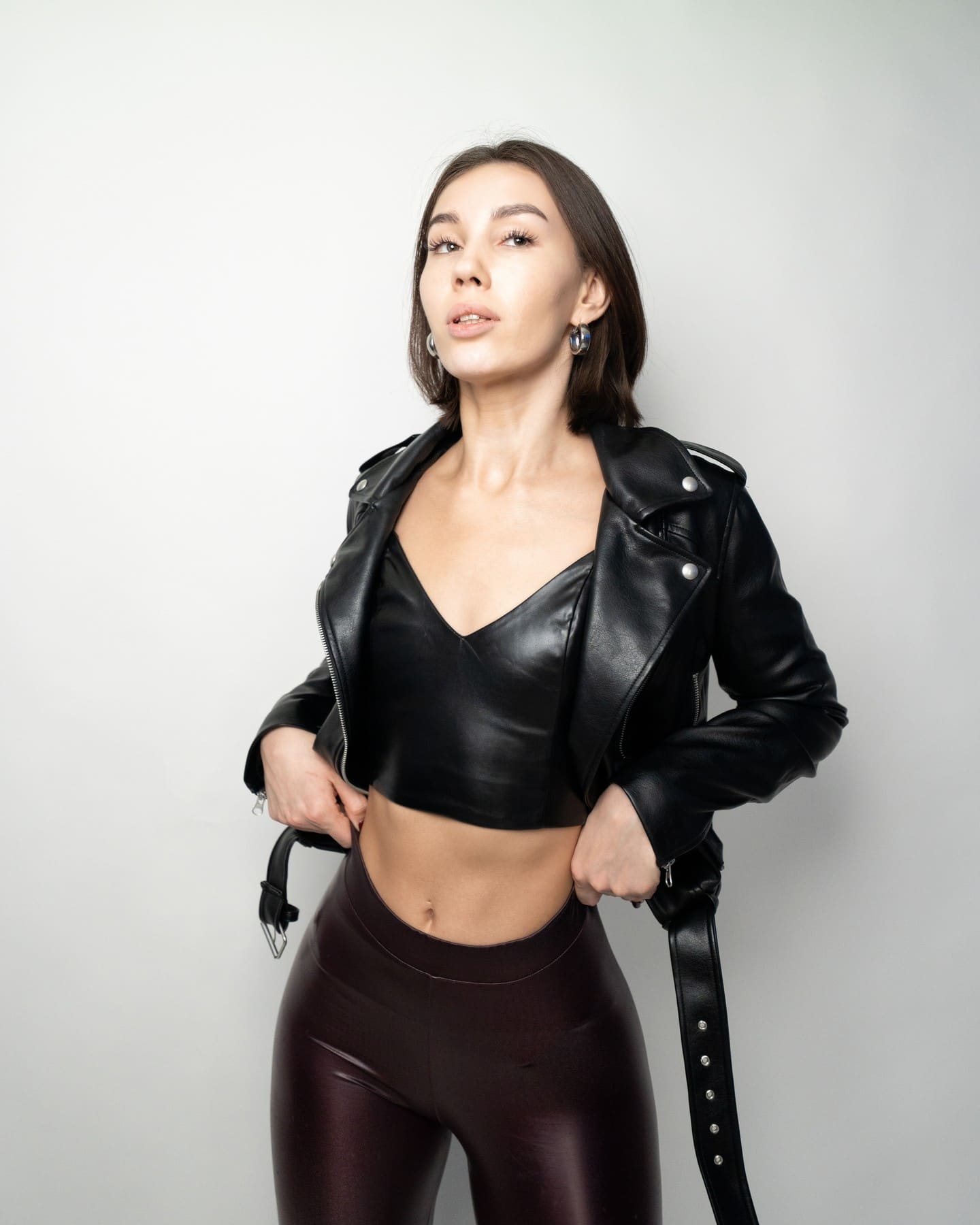 Sarah stands against a light background in a black leather jacket, cropped top, and dark leggings, resting her hands on her lapels.