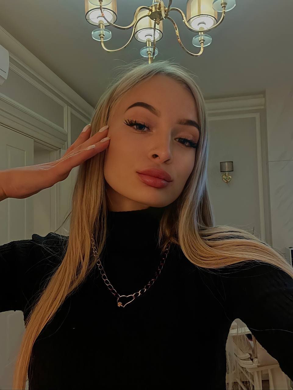 A blonde woman in a black turtleneck and chain necklace poses with her hand near her face in a room featuring a chandelier and paneled walls.
