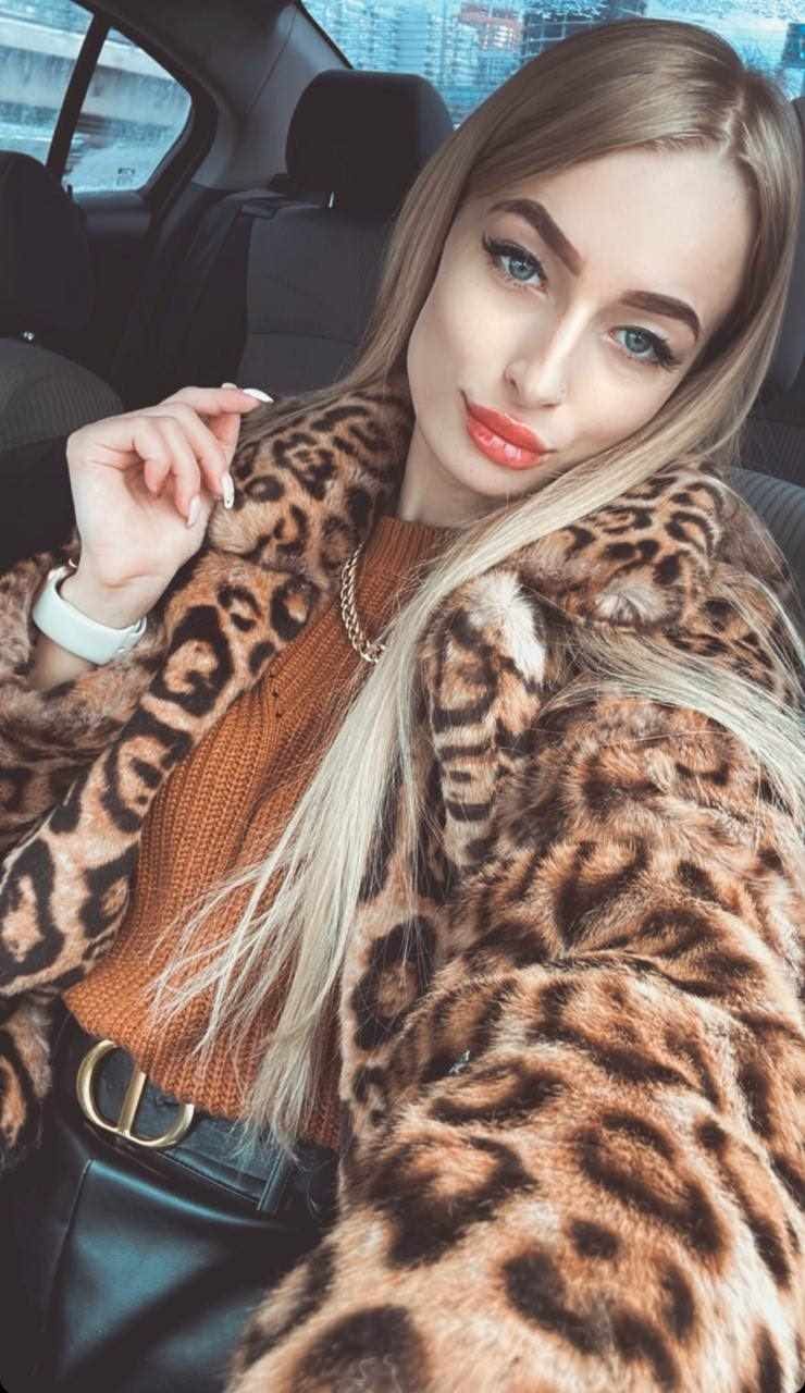 Kristina sits in a car back seat wearing a leopard-print coat, rust knit top, and black leather pants, her right hand raised near her shoulder.
