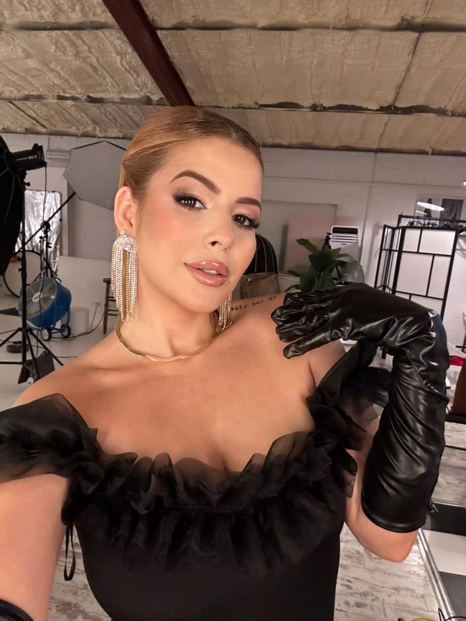 A woman with pulled-back blonde hair wears a black ruffled top and long black gloves, posing in a studio with lighting equipment visible.