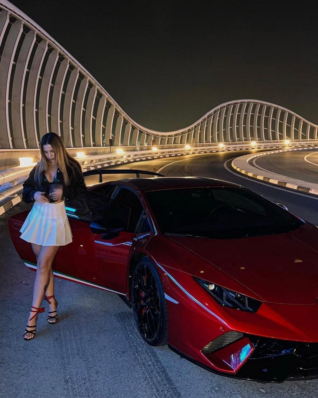 Woman stands by a red sports car at night wearing a black jacket, white pleated skirt, and high heels.