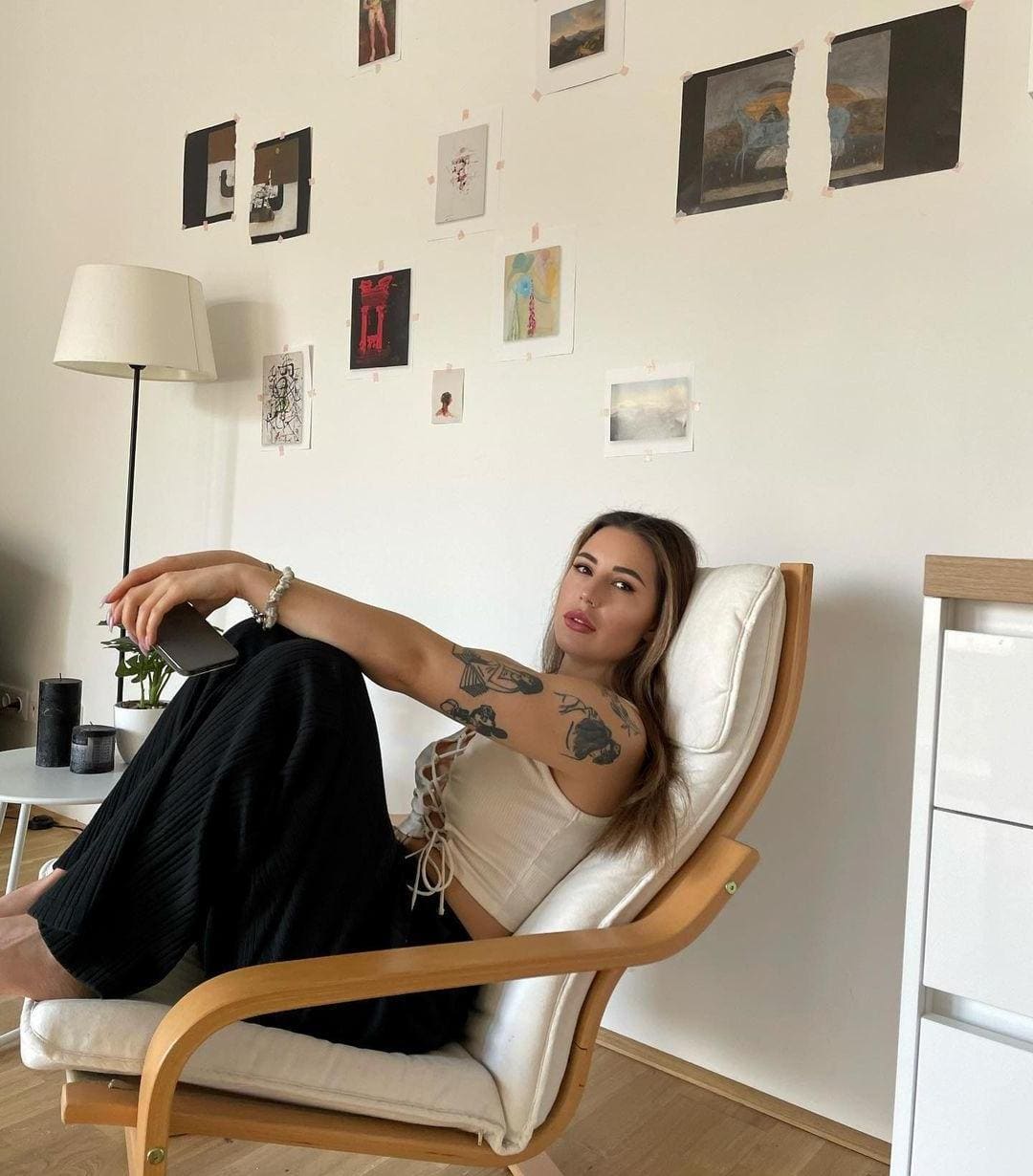 Woman with arm tattoos leans back in a wooden armchair wearing a white lace-up crop top and black pants.