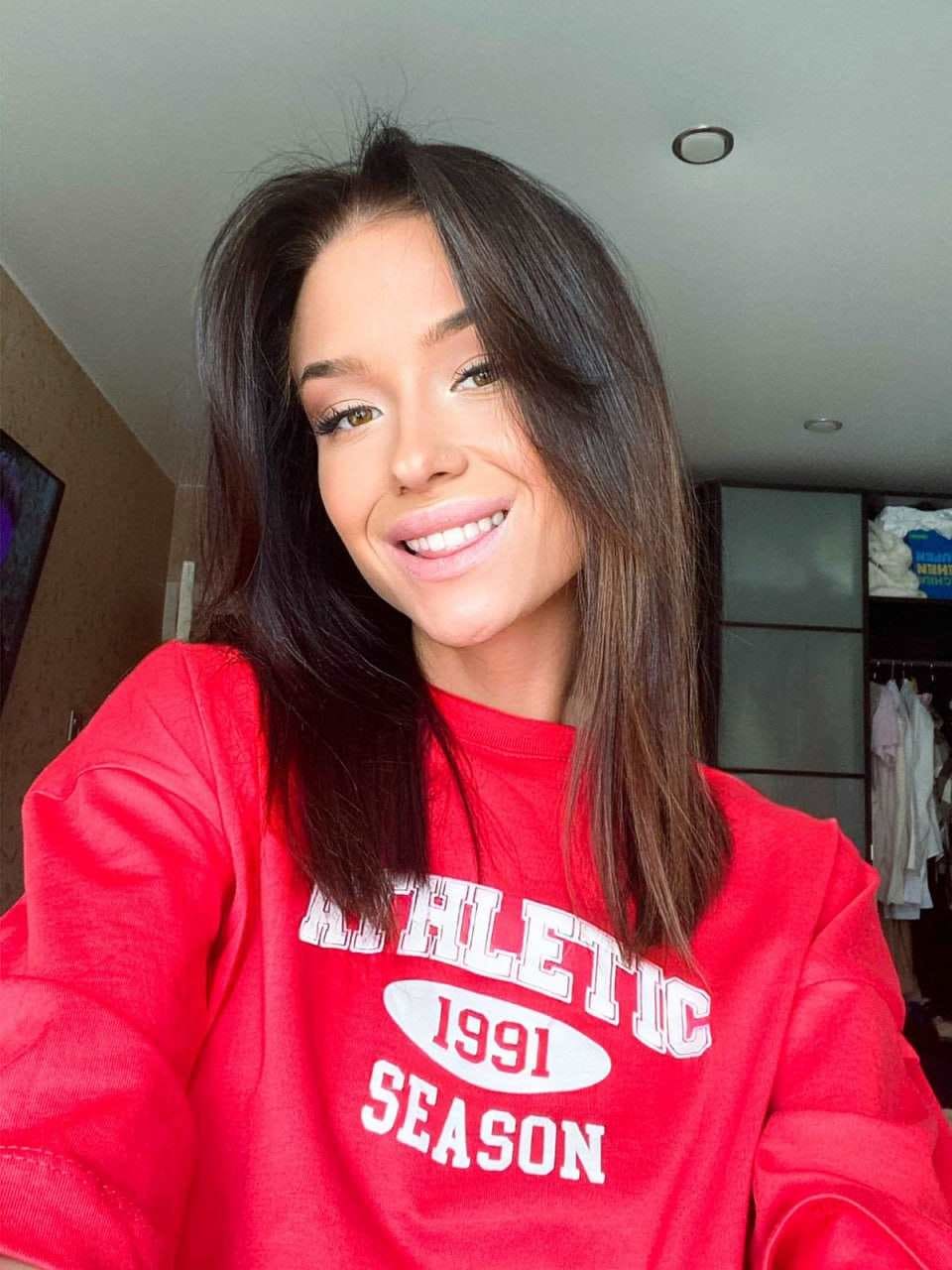 Lika smiles in a red sweatshirt with white lettering, seated indoors near a closet and wall art.