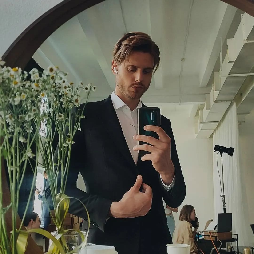 A man in a black suit jacket and open white shirt takes a mirror selfie, adjusting his jacket, with daisies and people in the background.