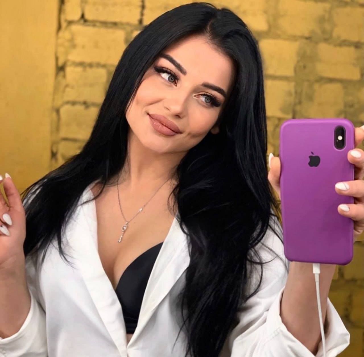 A woman takes a mirror selfie with a purple phone, wearing an open white shirt over a black bra against a yellow brick wall.