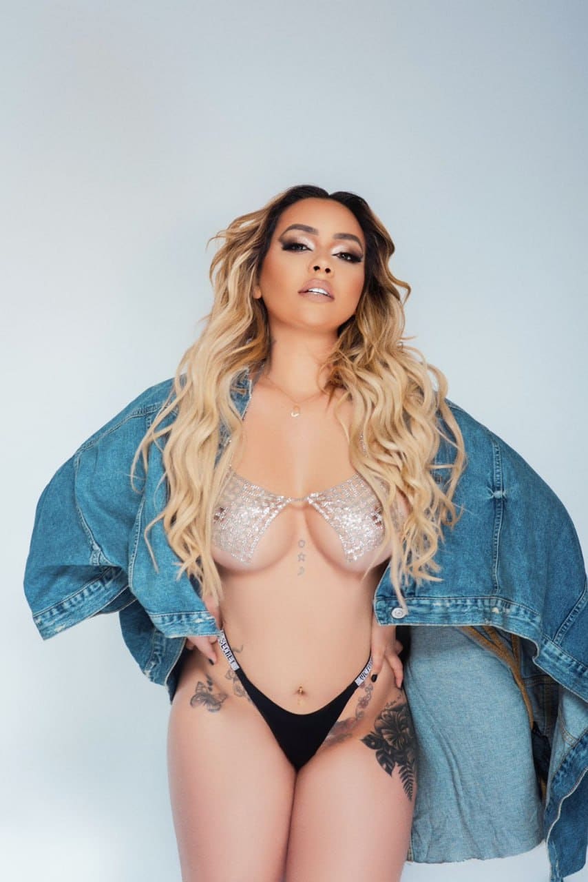A woman in a sparkly bra, black bottoms, and an off-shoulder denim jacket stands against a light background.