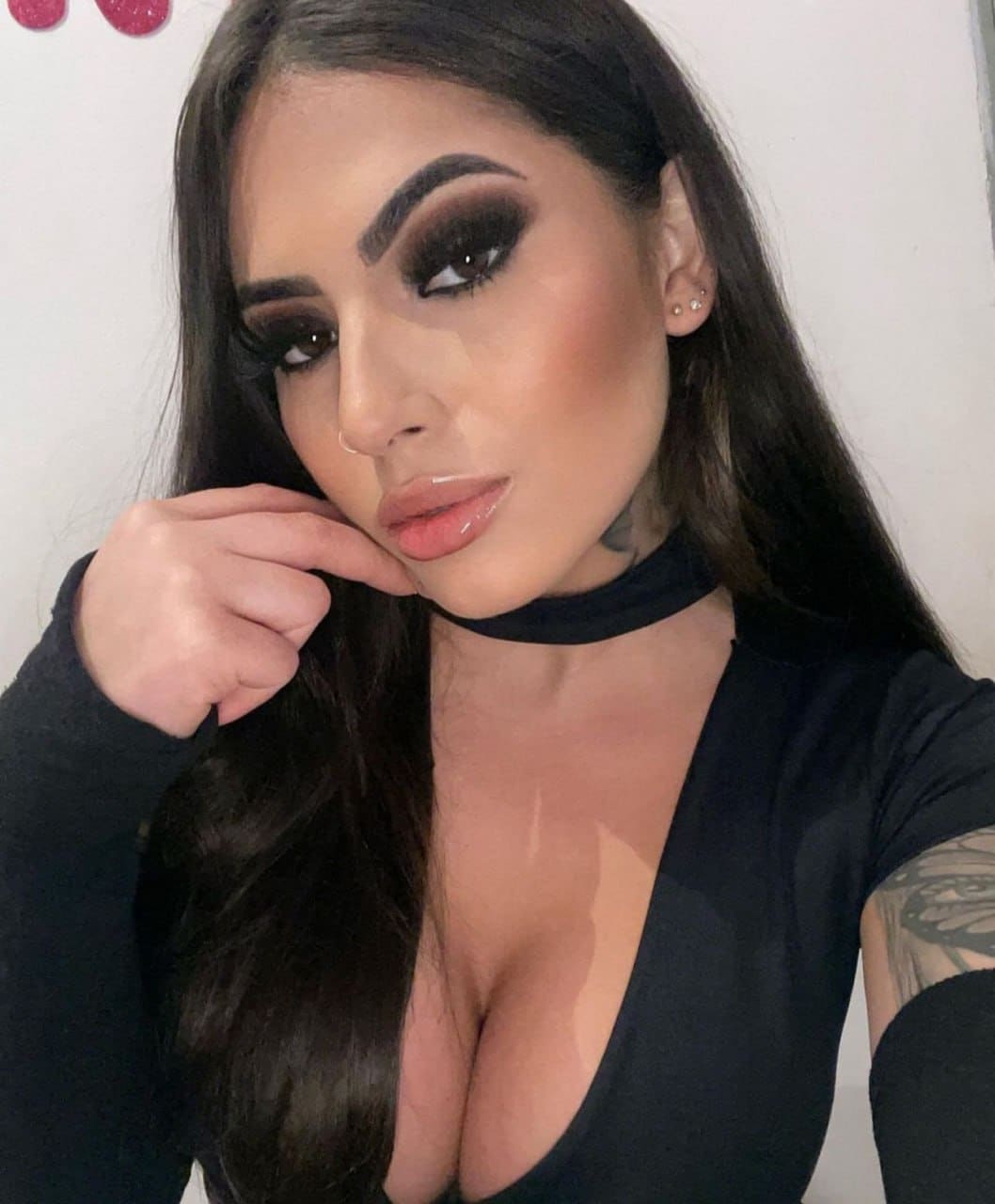 A woman with heavy eye makeup poses with a hand near her chin, wearing a black deep V-neck top and choker against a wall.