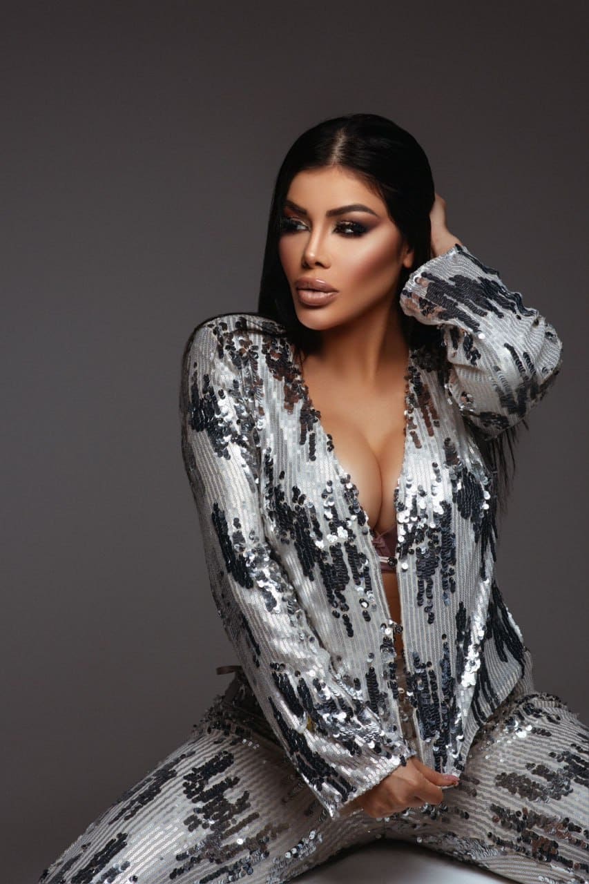 Woman in a silver and black sequined jacket and pants, seated in a studio with gray background, one hand raised to head, the other on her lap.