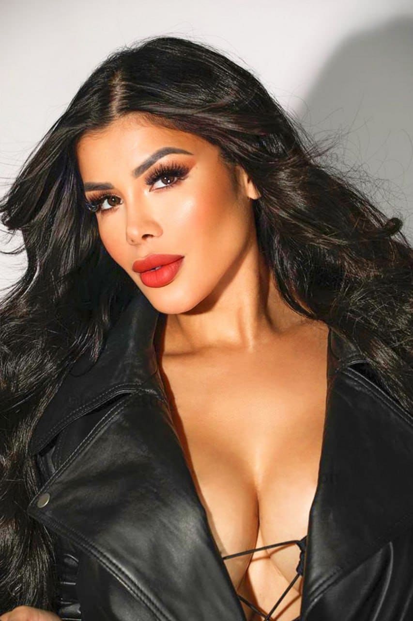 Camila with long dark wavy hair, wearing an open black leather jacket over a strappy top, shown from the chest up against a light background.
