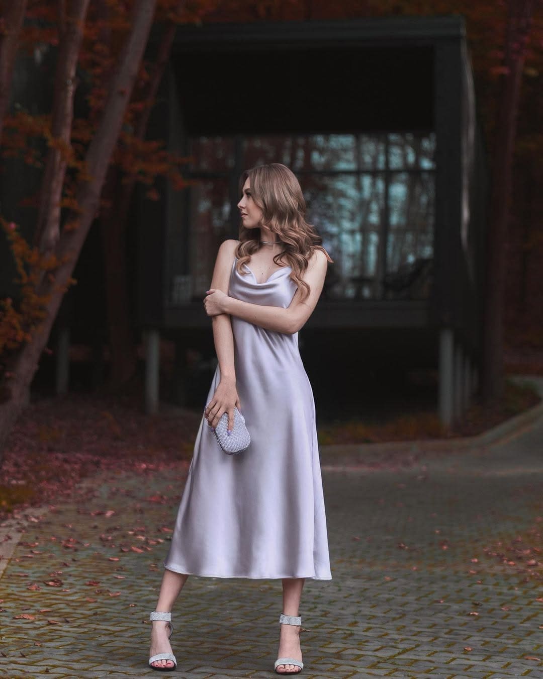 A woman in profile wears a silver satin midi dress and heels, holding a clutch on a cobblestone path near autumn trees.