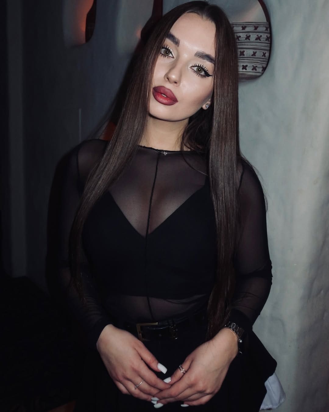 Stella stands facing forward in a black top with sheer mesh panels and dark pants, hands clasped, against a dim interior wall.