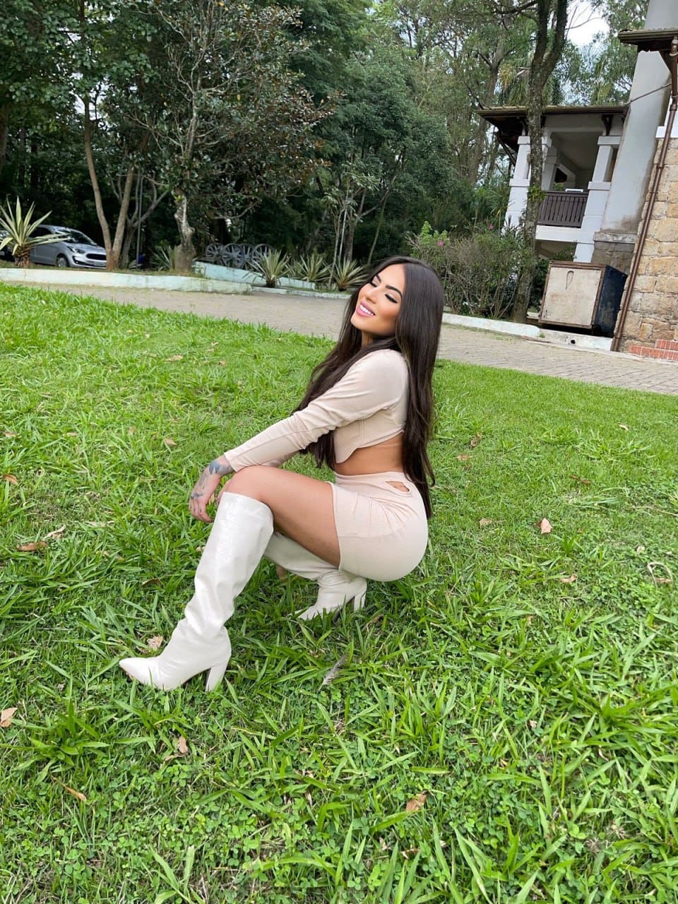 A woman crouches on a lawn in a beige crop top, mini skirt, and white knee-high heeled boots, with trees in background.