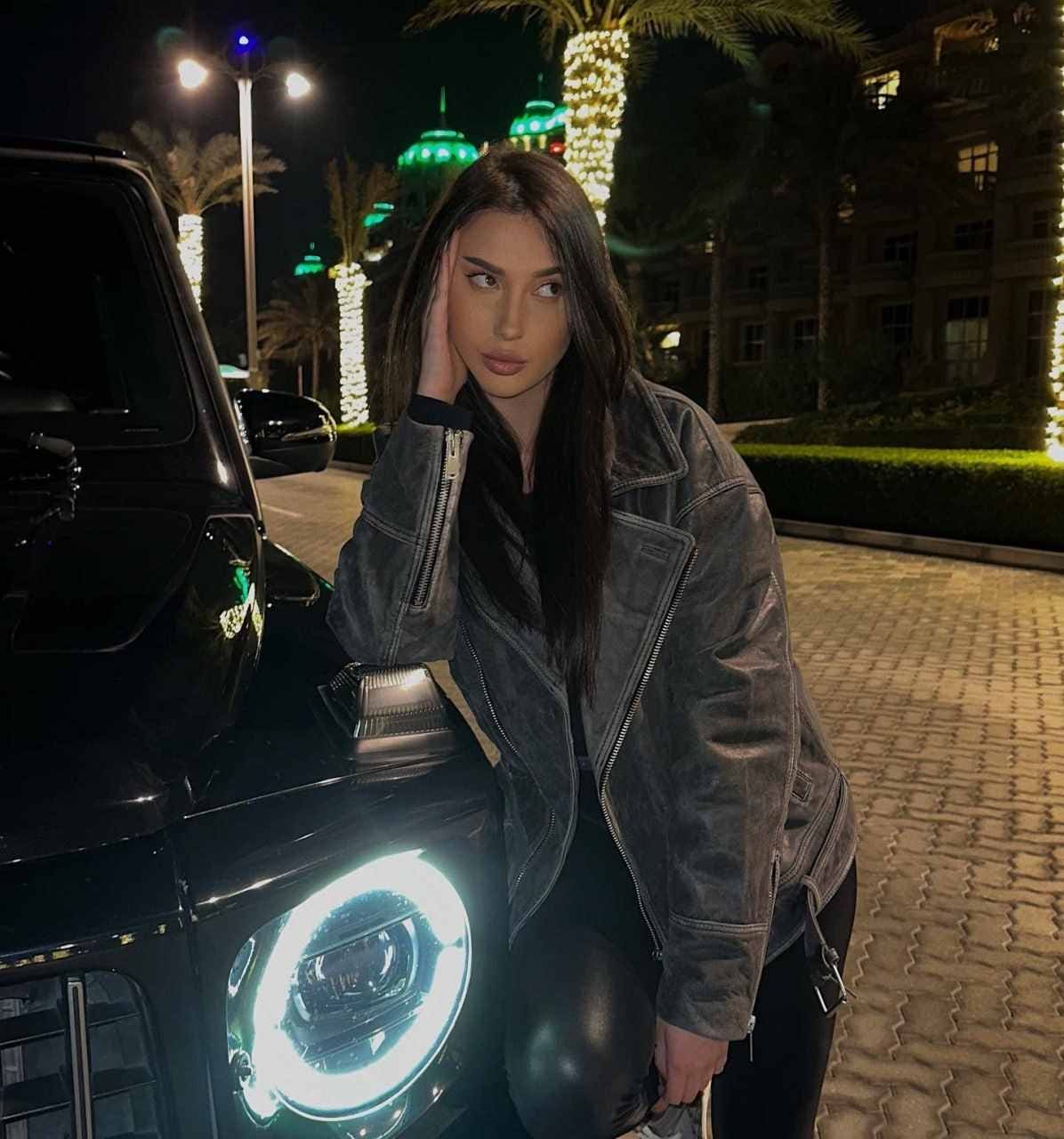 A woman in a grey leather jacket leans against a black car at night, hand on head, near illuminated palms.