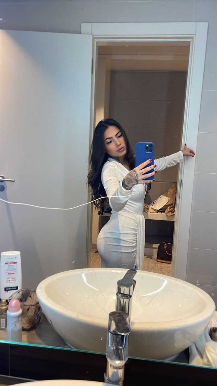 A mirror selfie shows a woman in a form-fitting white long-sleeved dress standing behind a bathroom vessel sink.