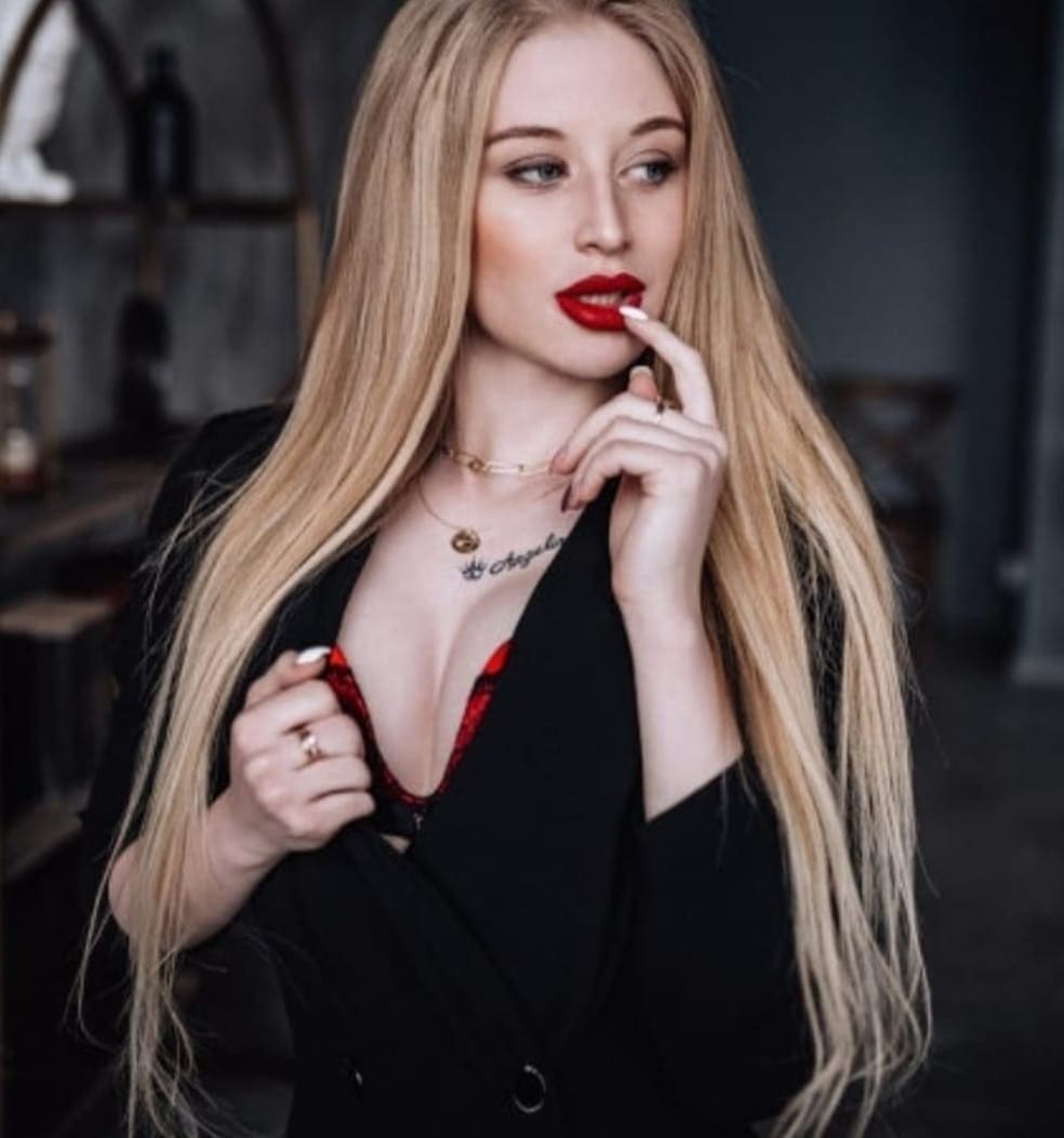 Angelina with long blonde hair wears a black blazer over a red lace top, posing indoors with one hand near her lips.