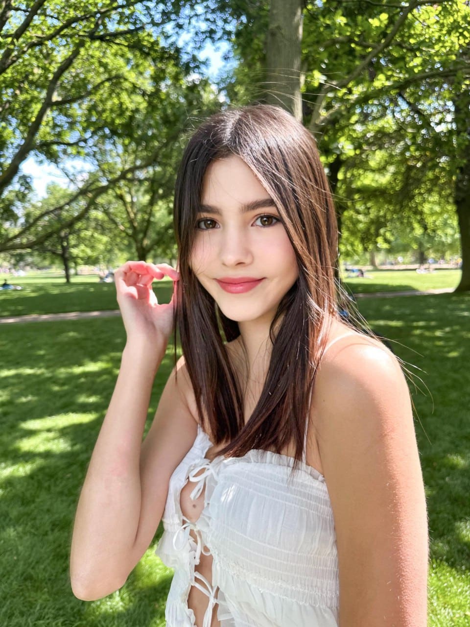 Calla stands in a sunlit park wearing a white lace-up top, her hand raised near her face as she looks toward the camera.