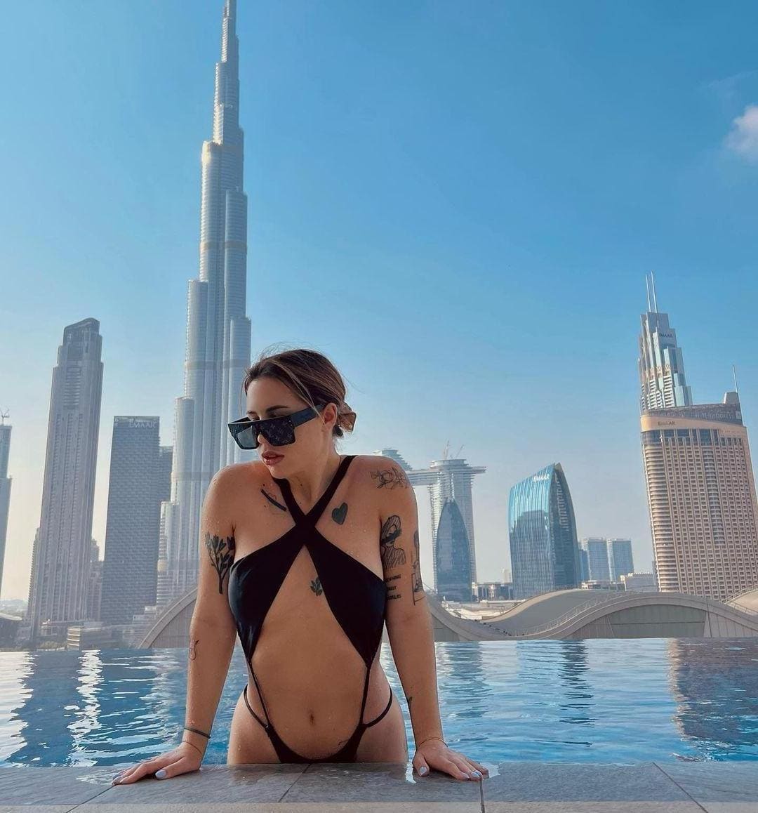 Woman in a black strappy swimsuit and sunglasses leans on a rooftop pool edge with a city skyline behind.