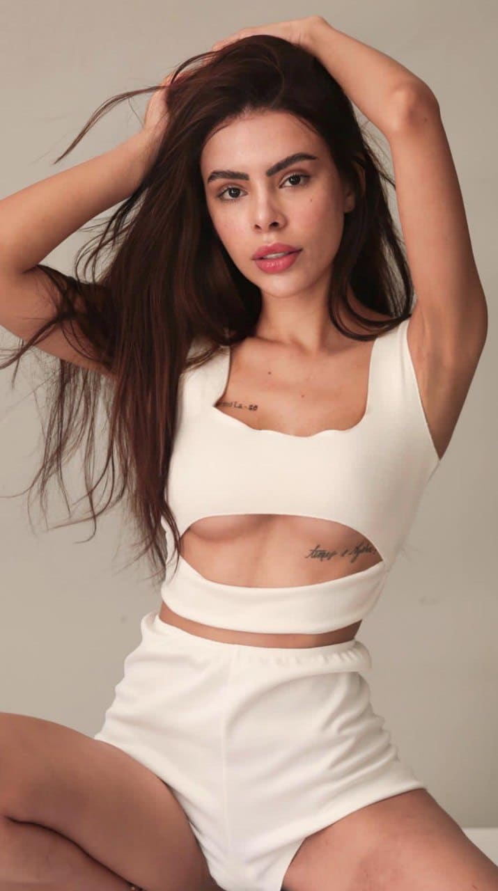 Ameline sits with hands in her long dark hair, wearing a white cut-out crop top and matching high-waisted shorts against a plain light background.