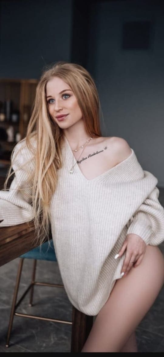A blonde woman sits on a stool in an oversized cream sweater slipped off one shoulder, revealing a tattoo and necklace.