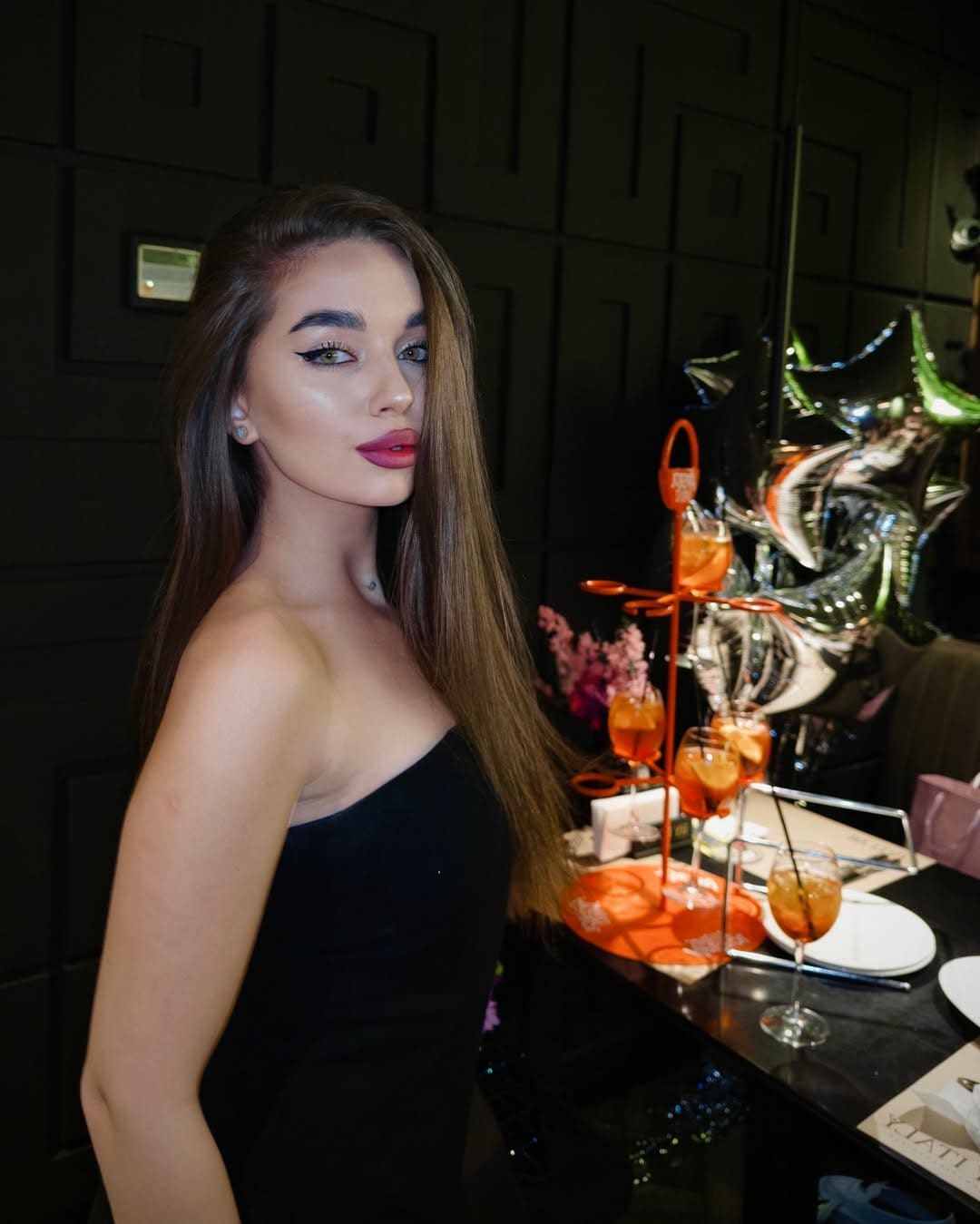 A woman in a black strapless top stands in a dim room, with orange drinks and silver star balloons on a table behind her.