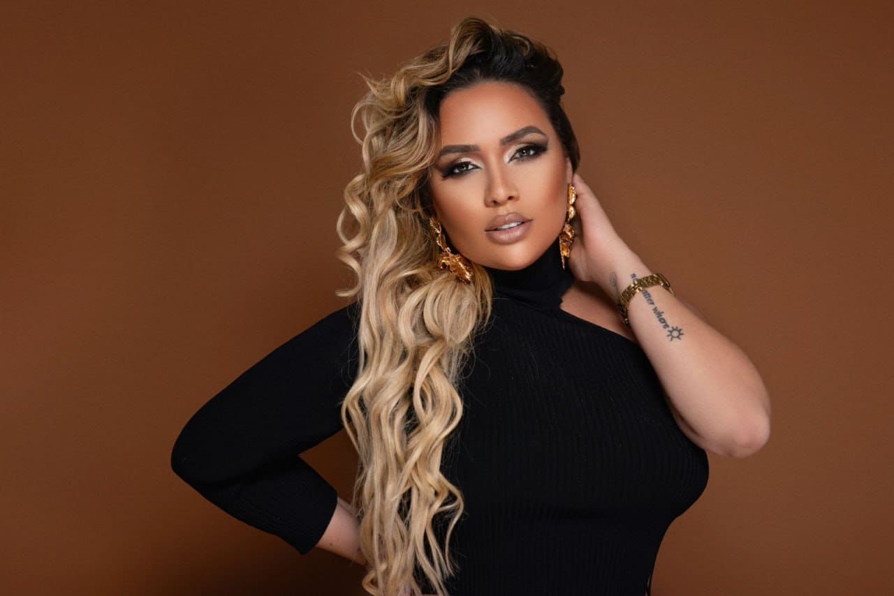 A woman in a black one-shoulder turtleneck and gold jewelry poses with a hand on her hip against brown.