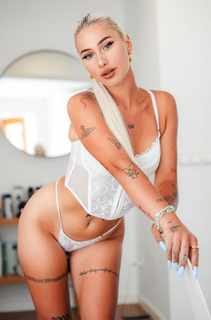 A blonde woman with a side braid and arm tattoos leans forward in a white lace bodysuit and briefs, with a round mirror on the wall behind her.