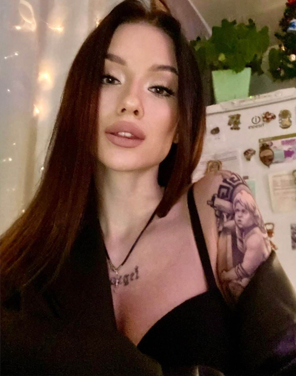A woman in a black low-cut top and dark jacket poses by string lights, showing a chest tattoo.