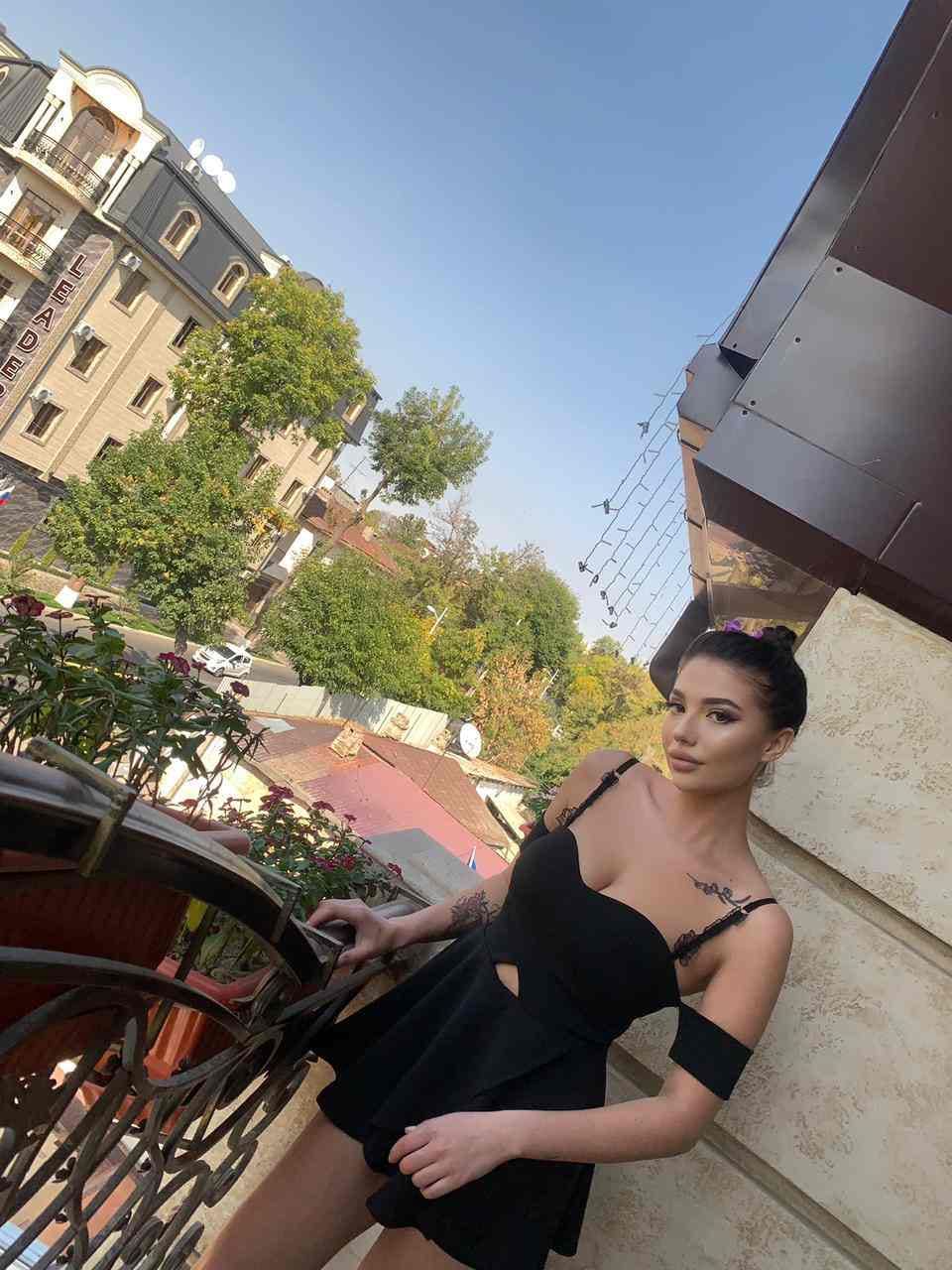 Woman in a short black dress leaning on a balcony railing, with buildings and trees visible under a clear sky.