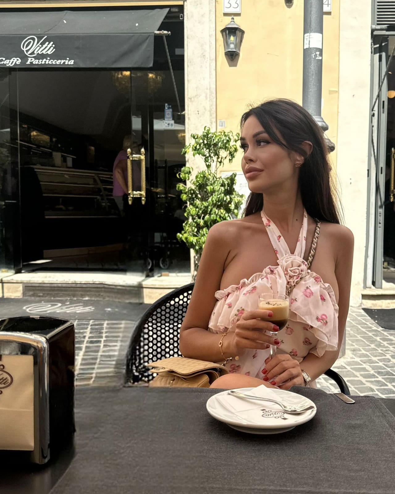 Sophia sits at an outdoor cafe table in a pink floral halter top, holding a coffee glass and looking to the side.