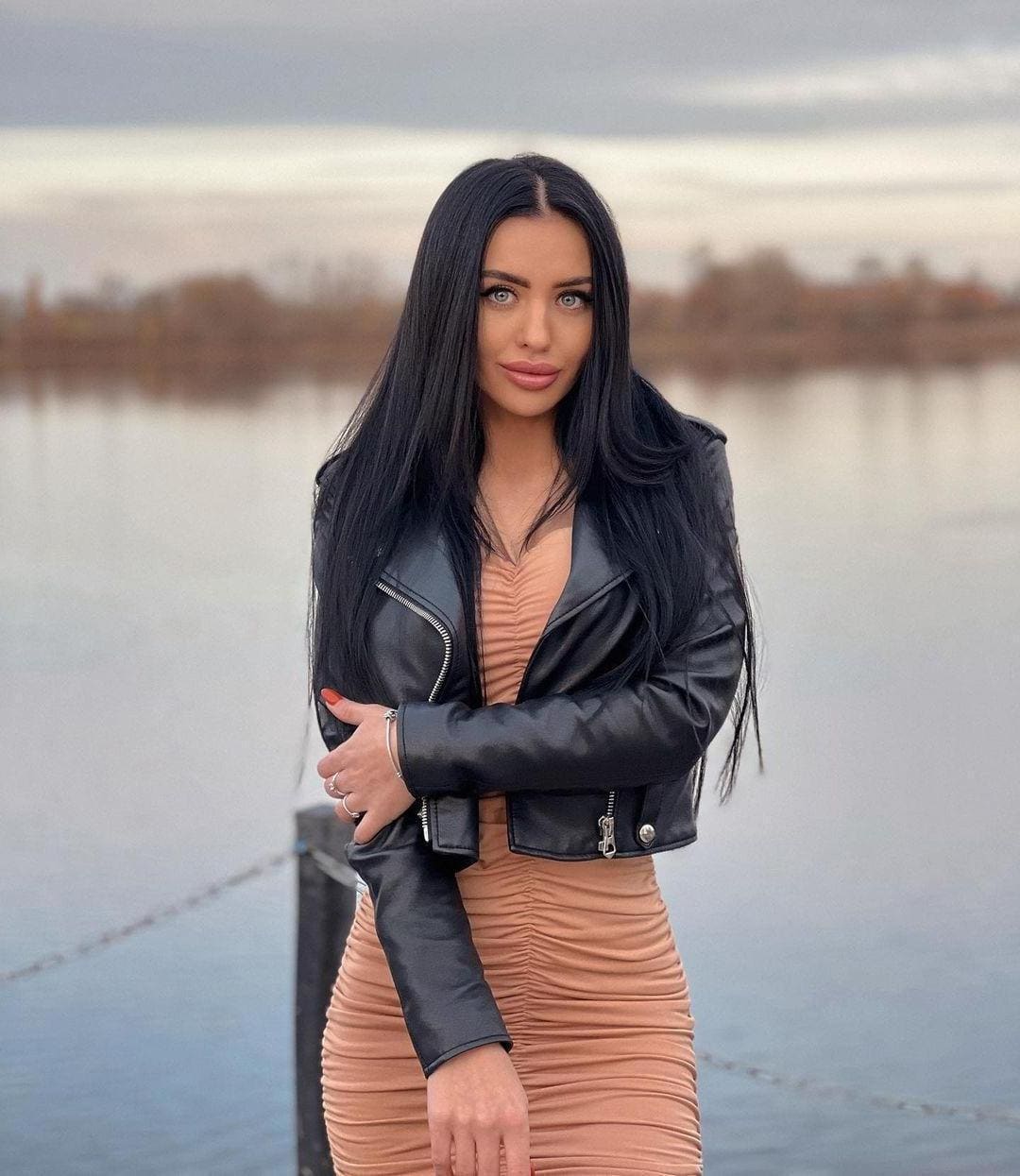 Camila stands with arms crossed in a black leather jacket and tan dress, against a calm body of water.