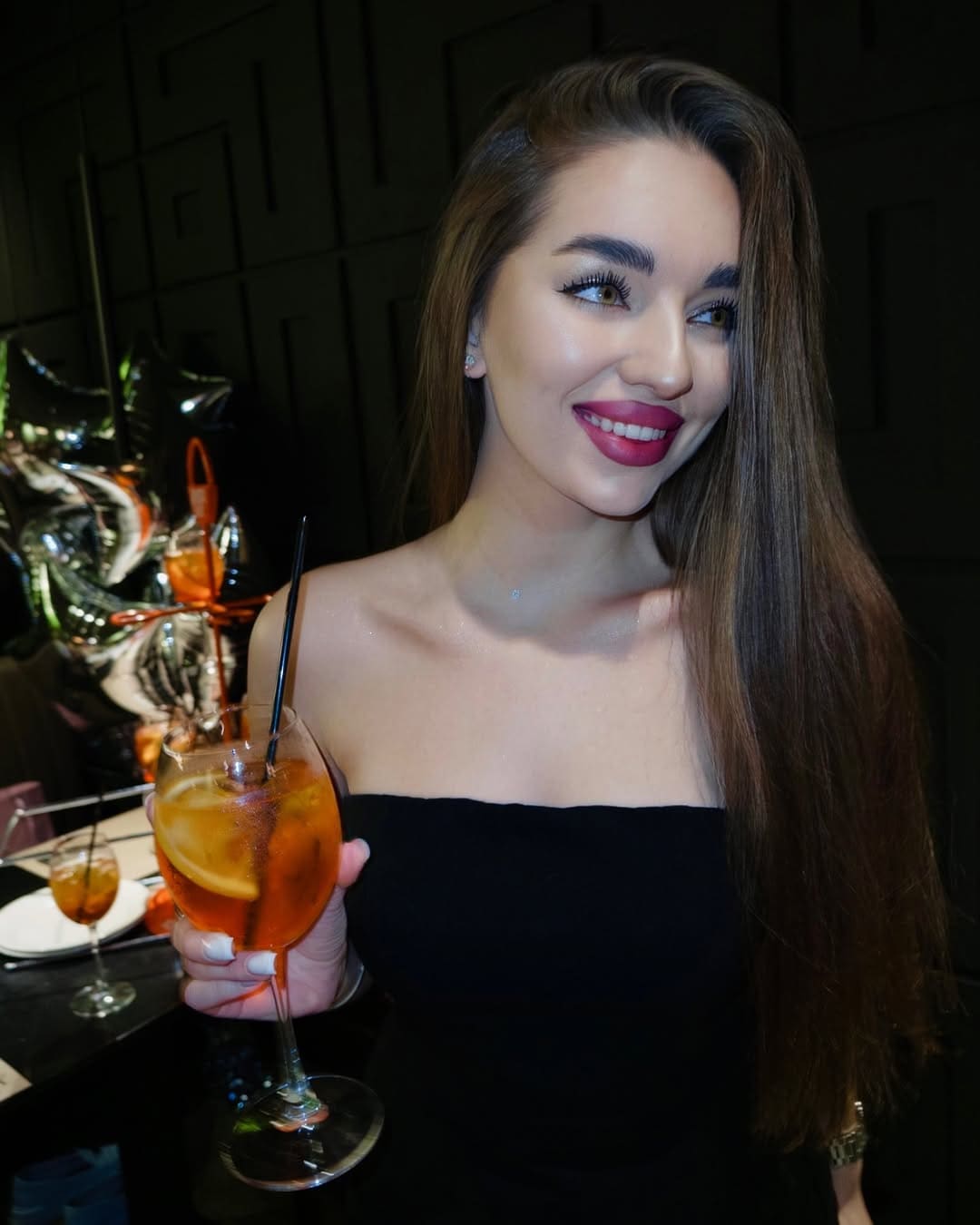 A woman in a black strapless top sits at a dark table holding a stemmed glass with an orange drink and a black straw.