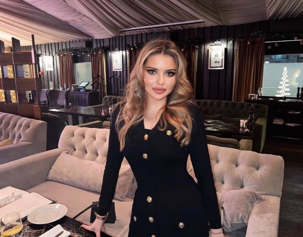 A woman in a long-sleeved black dress with gold buttons stands in a dim lounge, resting a hand on a table near a tufted sofa.