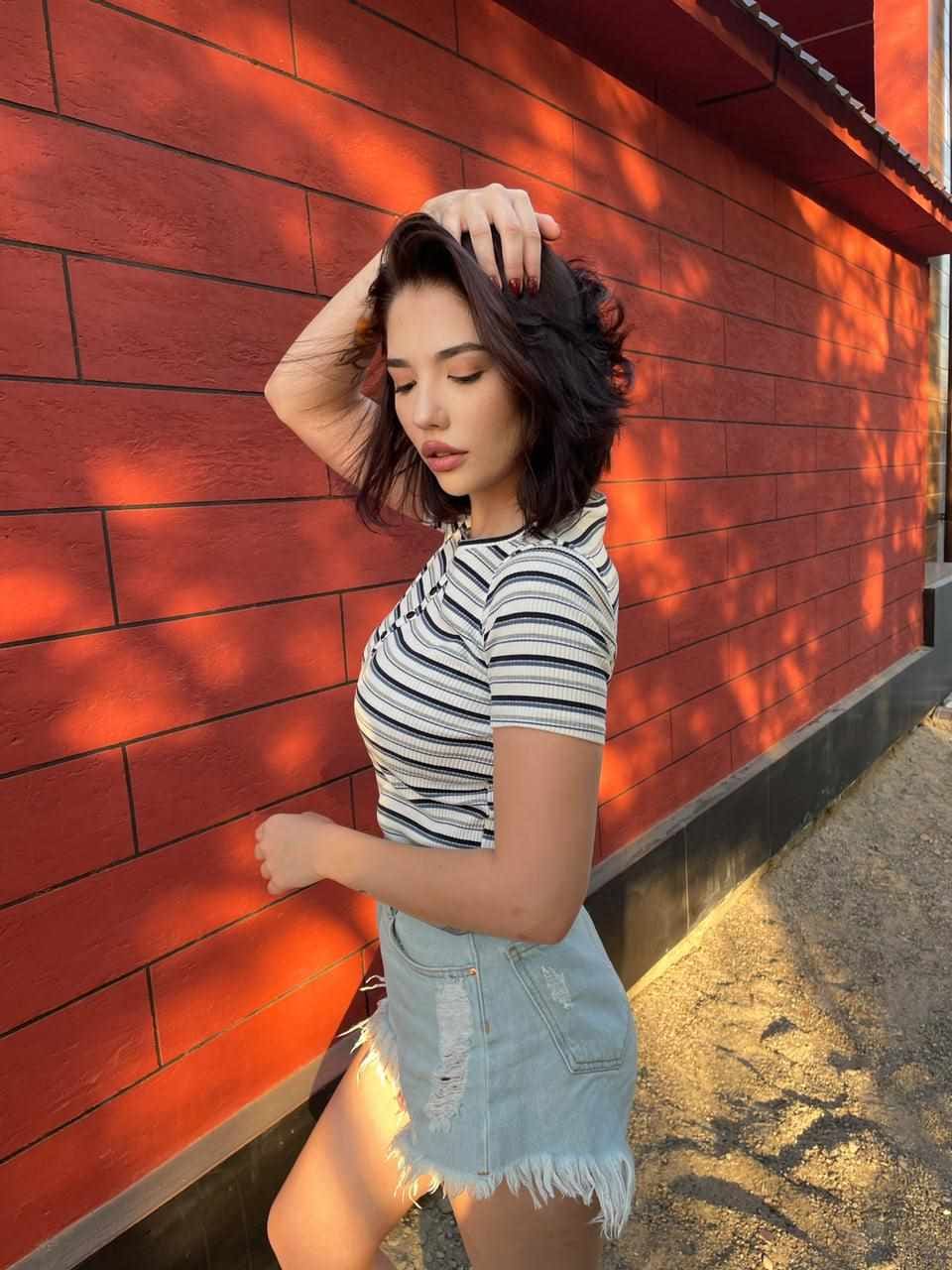 Woman in profile against a red brick wall, wearing a striped top and frayed denim mini skirt, one hand raised to her hair.