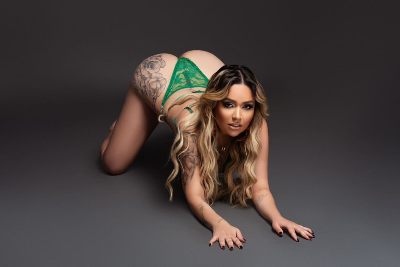 A woman in green lace lingerie kneels on a dark gray floor, showing tattoos on her hip and arms.