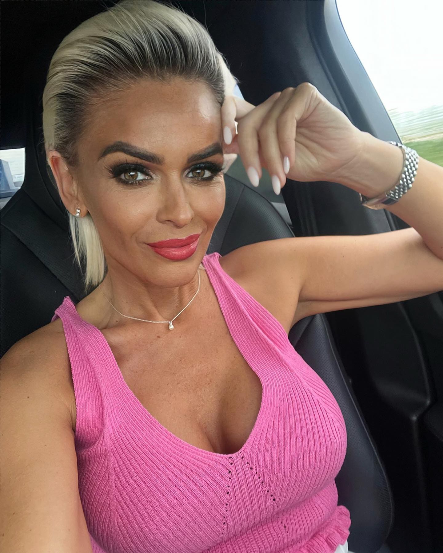 A woman with pulled-back blonde hair sits in a car wearing a pink ribbed sleeveless top and watch, raising one hand near her head.